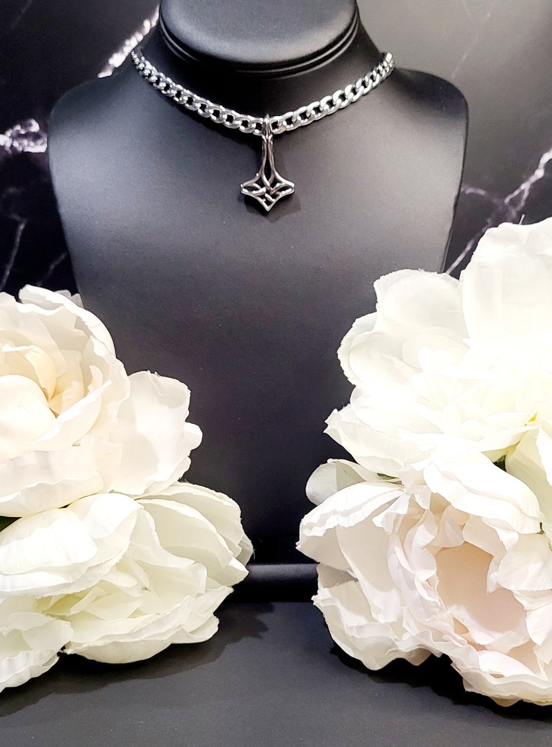 May include: A silver chain choker necklace with a geometric pendant. The necklace is on a black mannequin bust and there are white flowers on either side of the bust.