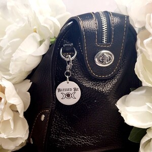 May include: A black leather handbag with a silver zip and a silver charm with the text "Blessed Be" and a triple moon symbol.