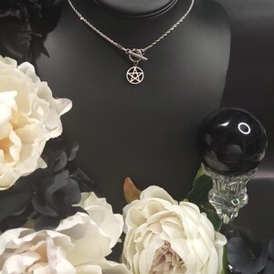 Op de afbeelding: Een zilveren ketting met een zilveren toggle-sluiting en een zilveren pentagram-hanger. De ketting wordt getoond op een zwarte mannequin-buste met witte bloemen op de achtergrond.