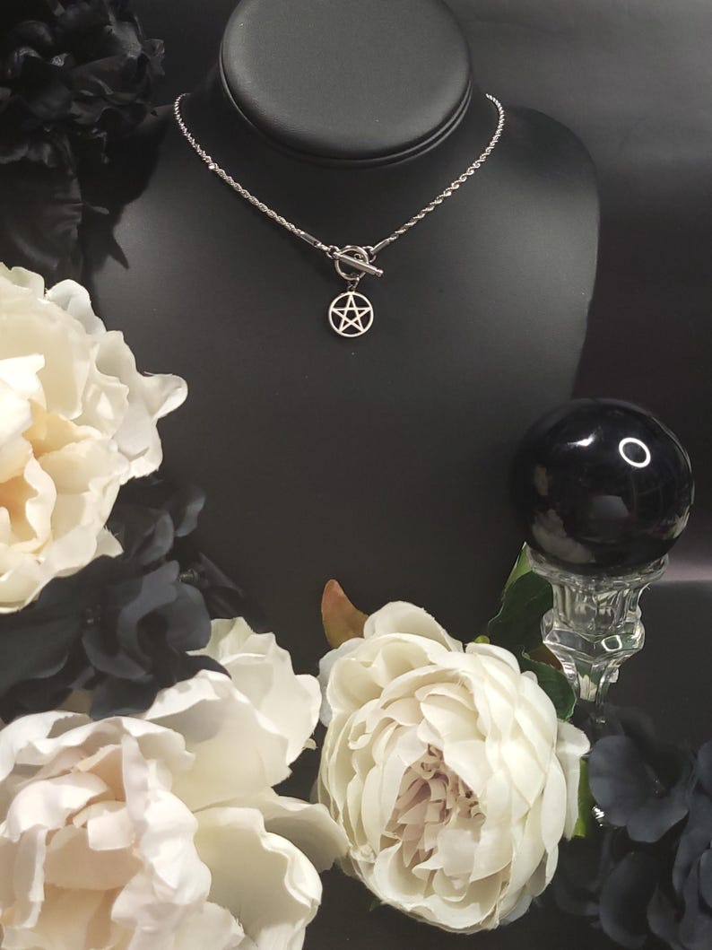 Op de afbeelding: Een zilveren ketting met een zilveren toggle-sluiting en een zilveren pentagram-hanger. De ketting wordt getoond op een zwarte mannequin-buste met witte bloemen op de achtergrond.