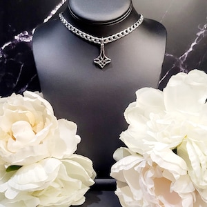 May include: A silver chain choker necklace with a geometric pendant. The necklace is displayed on a black mannequin bust with white flowers on either side.
