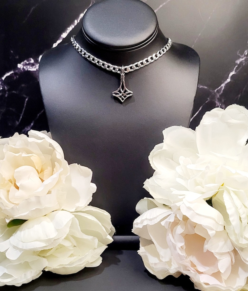 May include: A silver chain choker necklace with a geometric pendant. The necklace is displayed on a black mannequin bust with white flowers on either side.