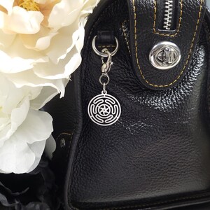 Puede incluir: Un charm plateado con un dise&ntilde;o circular que presenta una estrella en el centro, unido a un bolso de mano de cuero negro con una cremallera y un cierre plateados.