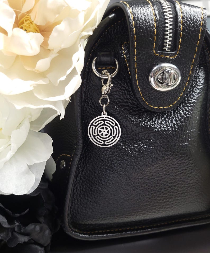 Puede incluir: Un charm plateado con un dise&ntilde;o circular que presenta una estrella en el centro, unido a un bolso de mano de cuero negro con una cremallera y un cierre plateados.