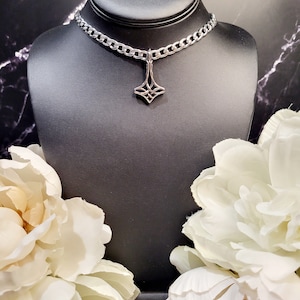 May include: A silver chain choker necklace with a silver pendant featuring a geometric design. The necklace is displayed on a black mannequin bust with white flowers on either side.