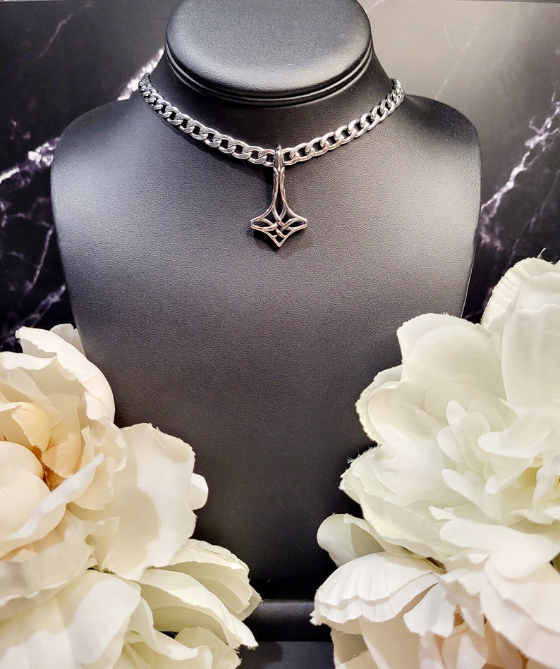May include: A silver chain choker necklace with a silver pendant featuring a geometric design. The necklace is displayed on a black mannequin bust with white flowers on either side.