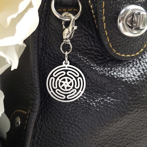 Puede incluir: Un charm plateado con un dise&ntilde;o circular que presenta una estrella y c&iacute;rculos conc&eacute;ntricos, unido a una cadena y un cierre plateados. El charm cuelga de un bolso de cuero negro.