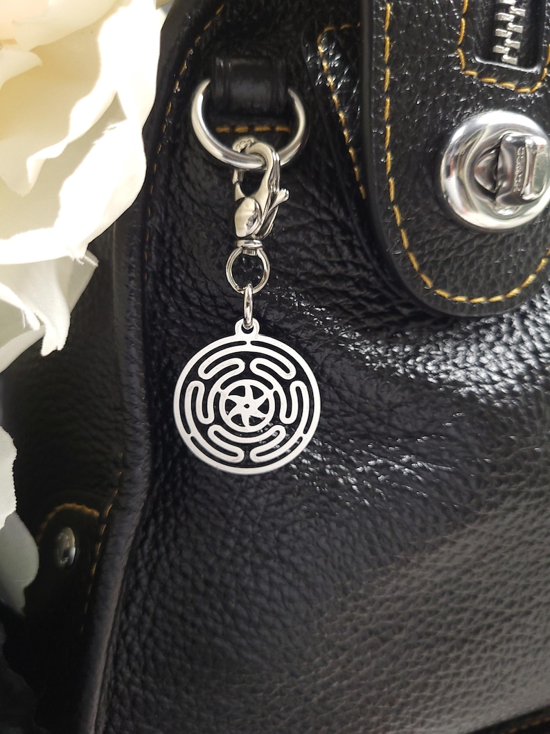 Puede incluir: Un charm plateado con un dise&ntilde;o circular que presenta una estrella y c&iacute;rculos conc&eacute;ntricos, unido a una cadena y un cierre plateados. El charm cuelga de un bolso de cuero negro.
