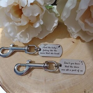 May include: Two silver keychains with engraved sayings. The top keychain says "And the lady's feeling like the moon that she loved." The bottom keychain says "Don't you know that the stars are all a part of us."