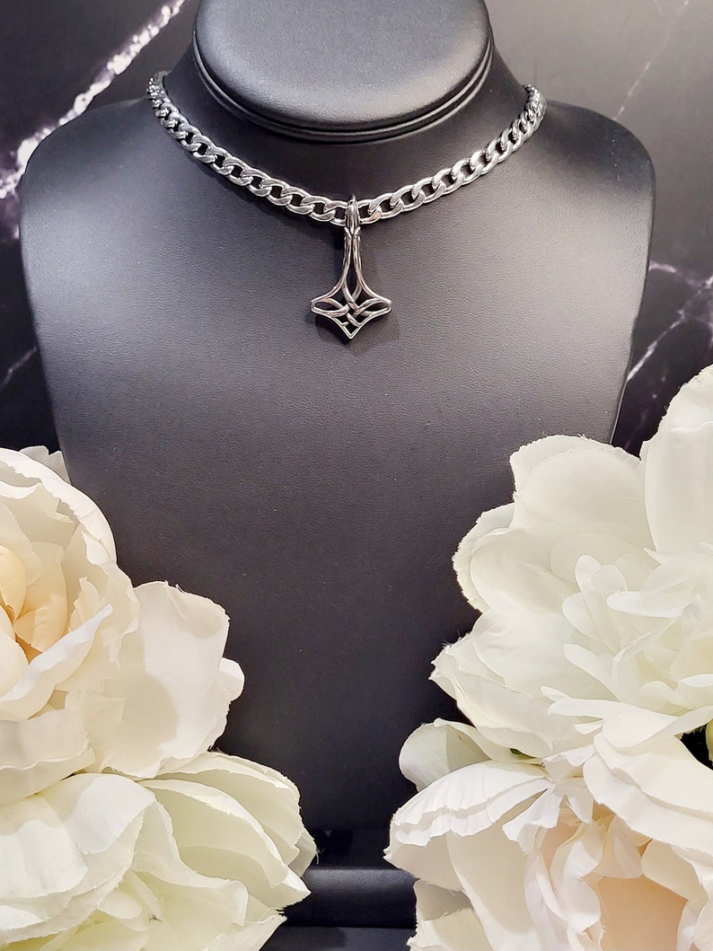 May include: A silver chain choker necklace with a silver pendant featuring a geometric design. The necklace is displayed on a black mannequin bust with white flowers on either side.