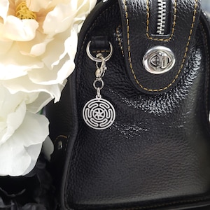 Puede incluir: Un bolso de mano de cuero negro con una cremallera plateada y un charm plateado sujeto al tirador de la cremallera. El charm es un dise&ntilde;o circular con una estrella en el centro.