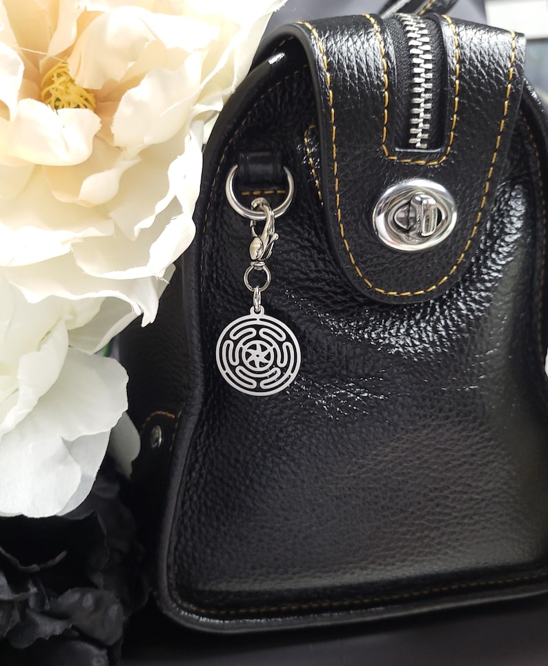 Puede incluir: Un bolso de mano de cuero negro con una cremallera plateada y un charm plateado sujeto al tirador de la cremallera. El charm es un dise&ntilde;o circular con una estrella en el centro.