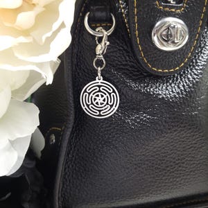 Puede incluir: Un charm plateado con un dise&ntilde;o circular cuelga de un cierre plateado unido a un bolso de mano de cuero negro.