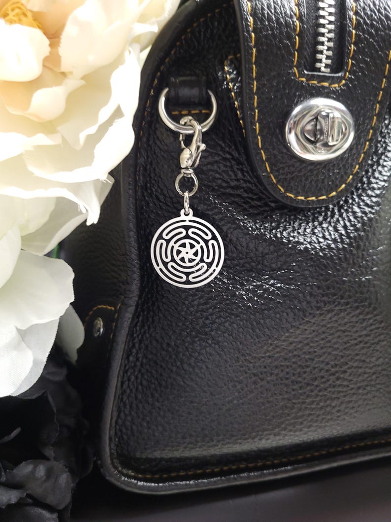 Puede incluir: Un charm plateado con un dise&ntilde;o circular cuelga de un cierre plateado unido a un bolso de mano de cuero negro.