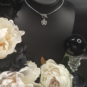 Op de afbeelding: Een zilveren ketting met een zilveren pentagram hanger en een toggle sluiting. De ketting wordt getoond op een zwarte mannequinbuste met witte bloemen op de achtergrond.