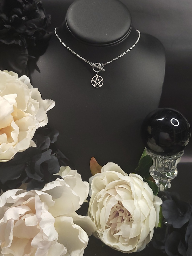 Op de afbeelding: Een zilveren ketting met een zilveren pentagram hanger en een toggle sluiting. De ketting wordt getoond op een zwarte mannequinbuste met witte bloemen op de achtergrond.