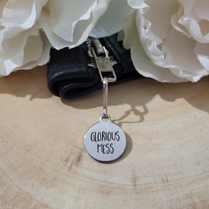 May include: A silver zipper pull with a round tag that says "Glorious Mess" in black lettering.