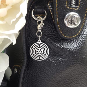 Puede incluir: Un charm plateado con un dise&ntilde;o circular que presenta una estrella y c&iacute;rculos conc&eacute;ntricos, unido a un bolso de mano de cuero negro con un cierre plateado.