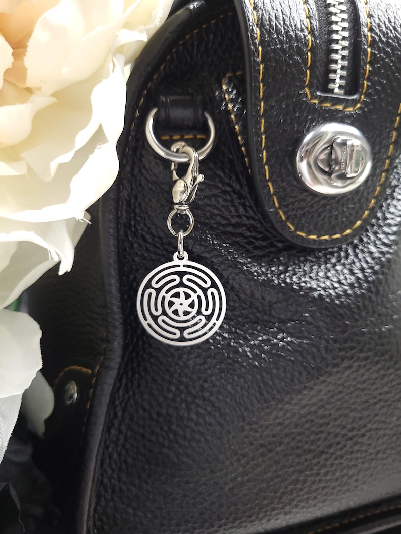 Puede incluir: Un charm plateado con un dise&ntilde;o circular que presenta una estrella y c&iacute;rculos conc&eacute;ntricos, unido a un bolso de mano de cuero negro con un cierre plateado.
