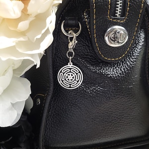 Puede incluir: Un bolso de mano de cuero negro con una cremallera plateada y un charm plateado sujeto al tirador de la cremallera. El charm es un dise&ntilde;o circular con una estrella en el centro.