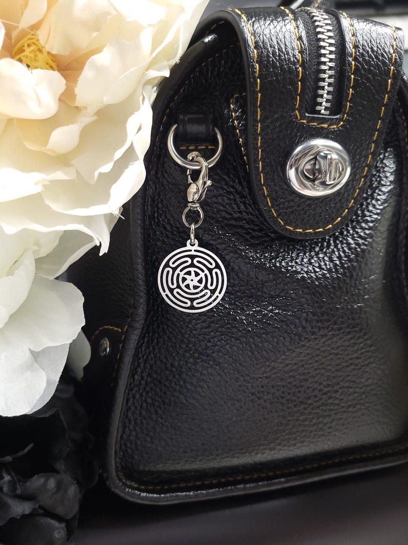 Puede incluir: Un bolso de mano de cuero negro con una cremallera plateada y un charm plateado sujeto al tirador de la cremallera. El charm es un dise&ntilde;o circular con una estrella en el centro.