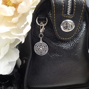 Puede incluir: Un bolso de mano de cuero negro con un dije plateado sujeto al tirador de la cremallera. El dije es un dise&ntilde;o circular con una estrella en el centro.