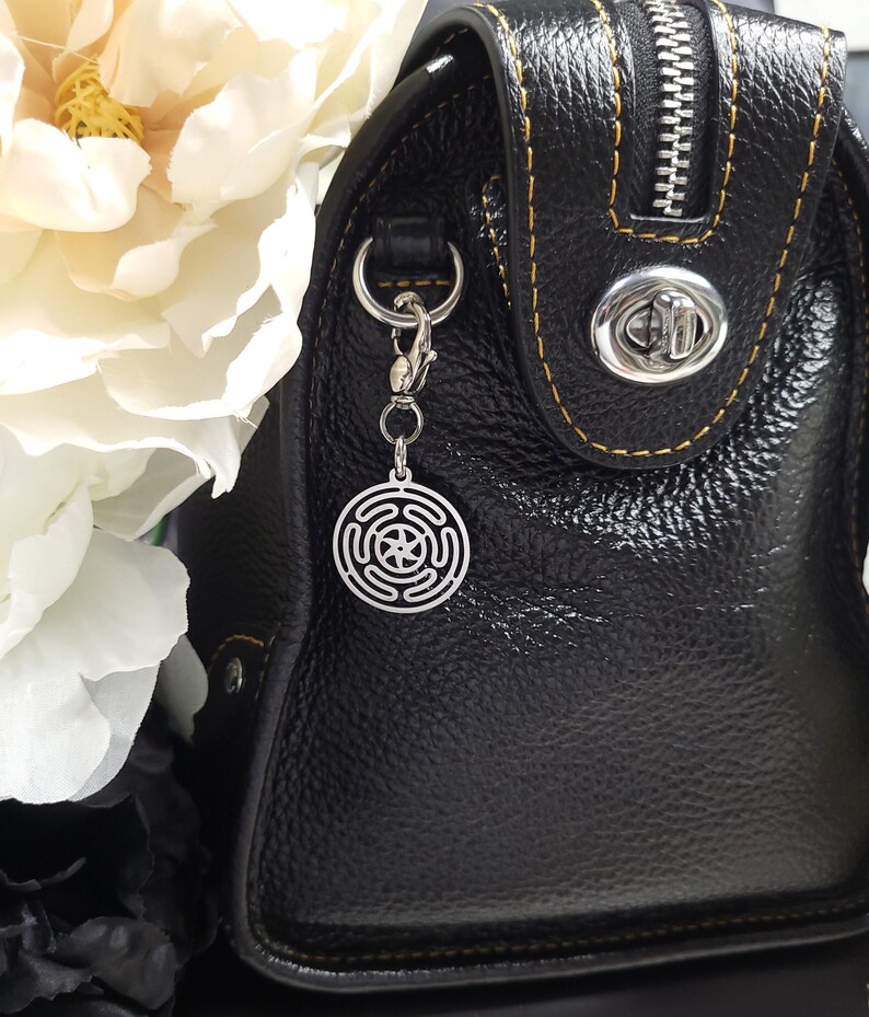 Puede incluir: Un bolso de mano de cuero negro con un dije plateado sujeto al tirador de la cremallera. El dije es un dise&ntilde;o circular con una estrella en el centro.