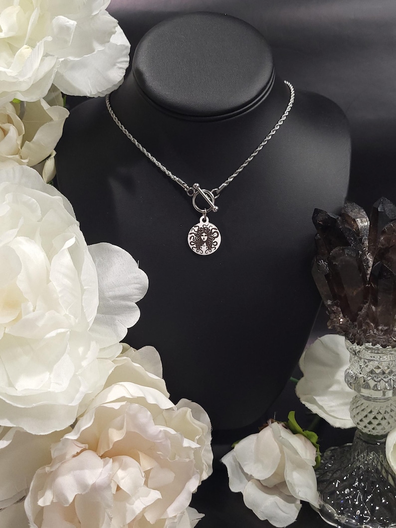 May include: A silver necklace with a toggle clasp and a round pendant featuring a detailed image of a woman's face with long hair.