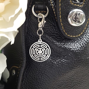Puede incluir: Un charm plateado con un dise&ntilde;o circular que presenta un patr&oacute;n geom&eacute;trico cuelga de un bolso de mano de cuero negro.