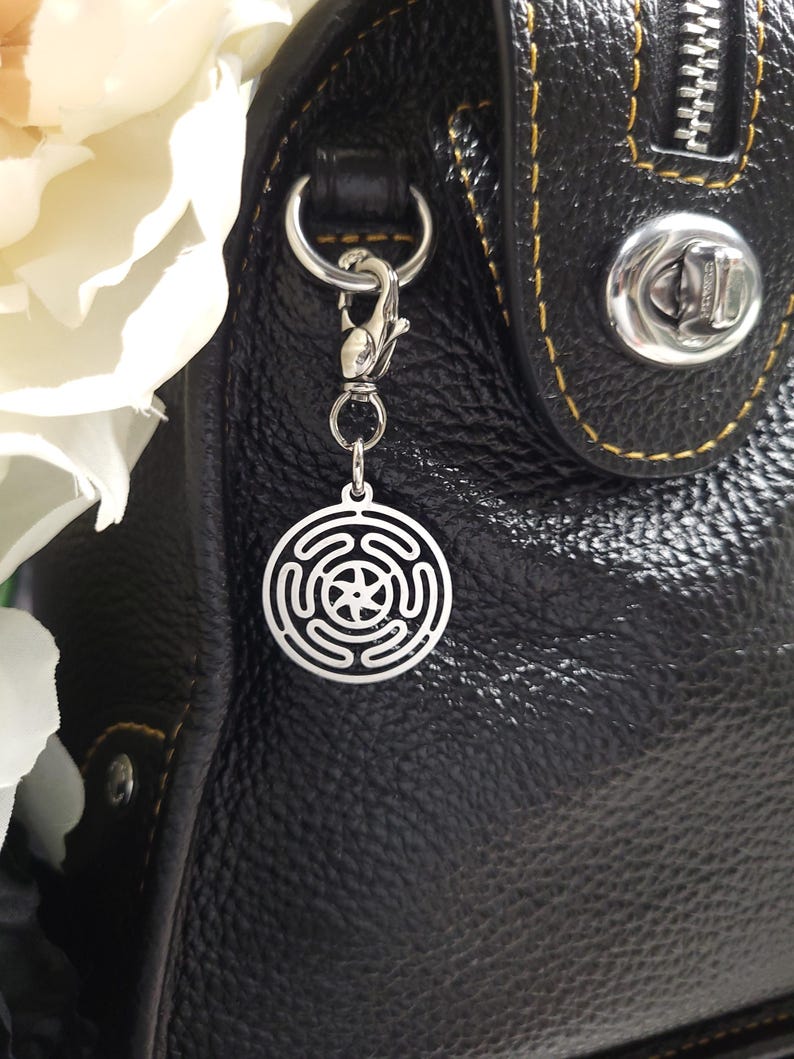 Puede incluir: Un charm plateado con un dise&ntilde;o circular que presenta un patr&oacute;n geom&eacute;trico cuelga de un bolso de mano de cuero negro.