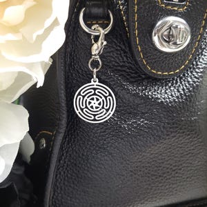 Puede incluir: Un charm plateado con un dise&ntilde;o circular que presenta una estrella de cinco puntas en el centro. El charm est&aacute; unido a un clip plateado y cuelga de un bolso de mano de cuero negro.