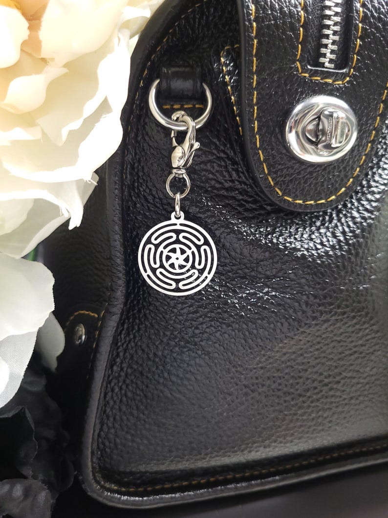 Puede incluir: Un charm plateado con un dise&ntilde;o circular que presenta una estrella de cinco puntas en el centro. El charm est&aacute; unido a un clip plateado y cuelga de un bolso de mano de cuero negro.