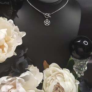 Op de afbeelding: Een zilveren ketting met een zilveren pentagram hanger en een toggle sluiting. De ketting wordt getoond op een zwart manikin met witte bloemen op de achtergrond.
