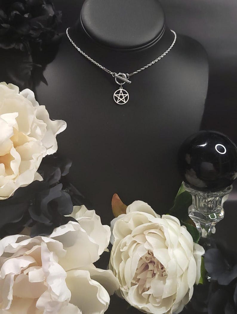 Op de afbeelding: Een zilveren ketting met een zilveren pentagram hanger en een toggle sluiting. De ketting wordt getoond op een zwart manikin met witte bloemen op de achtergrond.