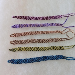 May include: Five handmade friendship bracelets in various colors. The bracelets feature a repeating diamond pattern. Colors include dark purple, light purple, brown, yellow and black, and blue. Each bracelet has braided ends.