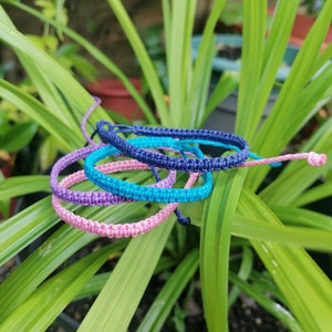 May include: A selection of macrame bracelets in shades of blue, turquoise, purple, and pink. The bracelets are displayed on green leaves, highlighting their vibrant colours and woven design. Each bracelet appears to be adjustable.