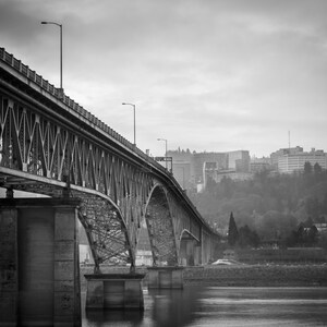 May include: A black and white photo of a steel bridge over a river. The bridge is made of steel beams and has a series of arches. The bridge is in the foreground and the city skyline is in the background.