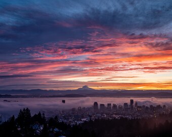May include: A scenic landscape featuring a city skyline silhouetted against a vibrant sunset. The sky is filled with streaks of pink, orange, and blue hues, with a mountain range and fog in the distance.