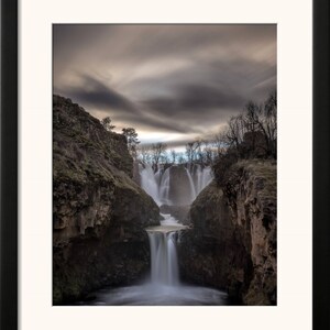 May include: A dramatic photograph of a waterfall cascading down a rocky cliff face. The water is flowing over multiple tiers, creating a misty spray. The sky above is a dark gray with wispy clouds.