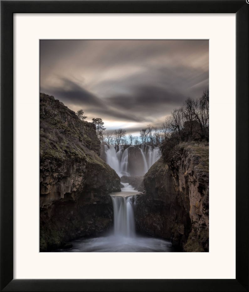 May include: A dramatic photograph of a waterfall cascading down a rocky cliff face. The water is flowing over multiple tiers, creating a misty spray. The sky above is a dark gray with wispy clouds.