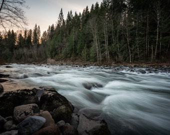 May include: A rushing river flows through a forest, with large rocks in the foreground and a line of trees in the background.