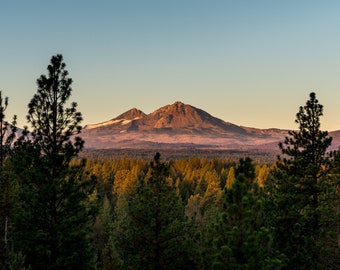 May include: A view of a mountain range with a forest in the foreground. The mountains are a reddish-brown color and the forest is a dark green. The sky is a light blue.