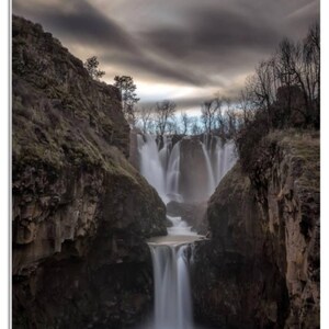 May include: A dramatic landscape photograph of a waterfall cascading down a rocky cliff face. The water is flowing over the edge of the cliff and down into a pool below. The sky is cloudy and overcast, and the trees are bare.