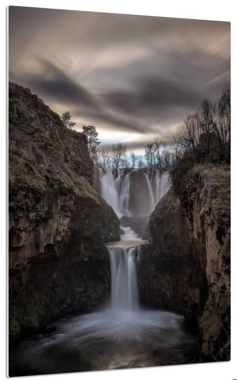 May include: A dramatic landscape photograph of a waterfall cascading down a rocky cliff face. The water is flowing over the edge of the cliff and down into a pool below. The sky is cloudy and overcast, and the trees are bare.