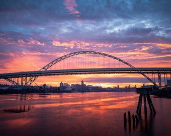 May include: A steel arch bridge over a river at sunset. The sky is a vibrant mix of pink, orange, and purple. The city skyline is visible in the distance.