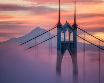 May include: A scenic photograph of a bridge shrouded in fog at sunset. The bridge's teal towers and cables are visible, with a mountain in the background. The sky is a vibrant mix of orange, pink, and purple hues.