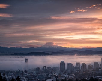 May include: A panoramic view of a city skyline with a mountain range in the background. The city is covered in fog, with a few skyscrapers peeking through. The sky is a vibrant orange and pink, with clouds streaking across the horizon.