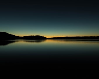 May include: A dark silhouette of a forest line reflected in a still lake at dusk. The sky is a gradient of blue and orange.