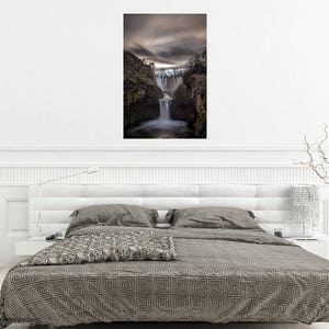 May include: A photograph of a waterfall cascading down a rocky cliff face. The waterfall is surrounded by trees and the sky is overcast. The image is in a minimalist white frame and is hanging on a white wall above a gray bed with a patterned duvet.
