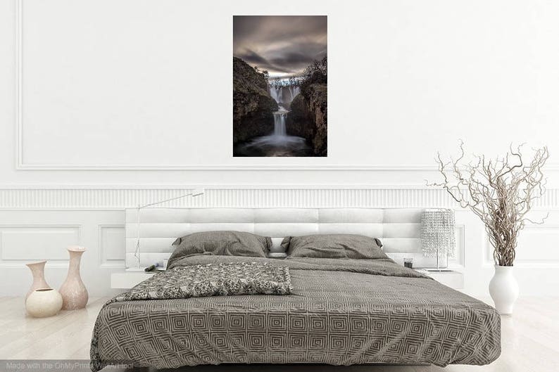 May include: A photograph of a waterfall cascading down a rocky cliff face. The waterfall is surrounded by trees and the sky is overcast. The image is in a minimalist white frame and is hanging on a white wall above a gray bed with a patterned duvet.